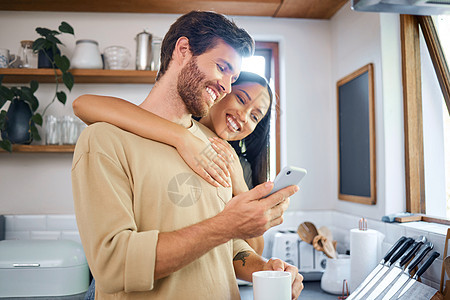 快乐的年轻跨种族夫妇在家里充满爱意和深情 年轻人用他的智能手机拿着咖啡杯 而他的女朋友从后面拥抱他 浏览社交媒体或发送短信男人妻图片