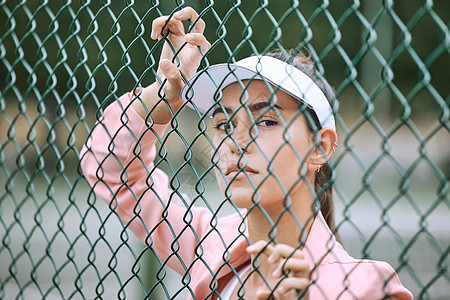 一名女运动员靠在铁丝网上的特写镜头 身穿白色遮阳板和粉色夹克的年轻西班牙裔网球运动员在网球场上摆姿势图片