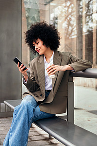 年轻妇女在等公车时使用她的电话长椅城市生活女性公共汽车企业家成人工人咖啡杯阅读电车背景