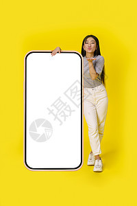 发送飞吻亚洲女孩站在巨大的智能手机旁边 智能手机上有白色屏幕 穿着黄色背景的休闲装 免费空间模拟移动应用程序广告图片