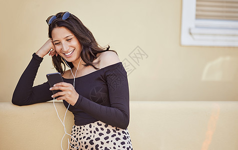 穿着耳机 在智能手机上监听音乐或观看滑稽视频的无忧心的年轻女子 漂亮女人站在人行道上时使用移动应用程序图片