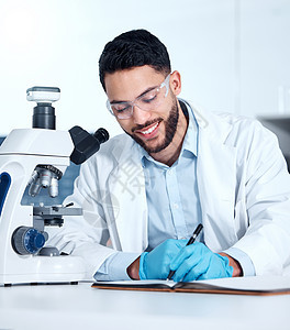 一位英俊的年轻混血男子戴着手套和实验室大衣 在实验室的显微镜下观察医学样本 一位戴着安全护目镜并在笔记本上写作时微笑的男性科学家图片