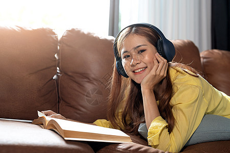 快乐的女孩听音乐和阅读书 躺在家里的沙发上图片