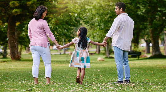 在阳光明媚的日子里 父母和小女孩在公园散步时手牵着手的后视图 有一个孩子的充满爱心和关怀的家庭 在户外玩得开心图片