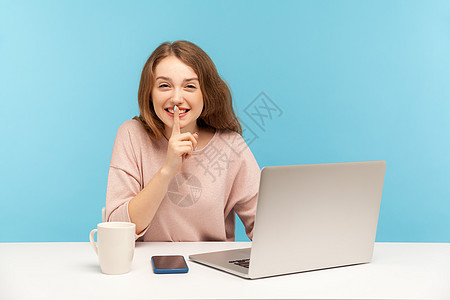 情感年轻女性在蓝背景的笔记本电脑上工作女士耳语人士微笑手指手势嘴唇工人互联网阴谋图片