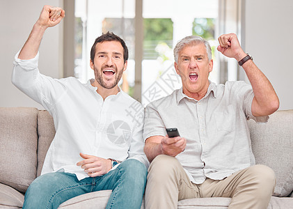 剪裁一个英俊的年轻男子和他年长父亲的肖像 在看家里沙发上的体育比赛时欢呼他们的团队图片