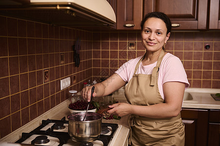 迷人的多民族家庭主妇微笑着看着相机 在生锈的家庭厨房准备自制樱桃果酱图片