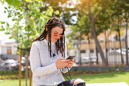 年轻女性嬉皮士 在粉红色太阳镜中戴了短锁 使用智能手机坐在露天的夏令营 新一代人 技术掌握在手图片