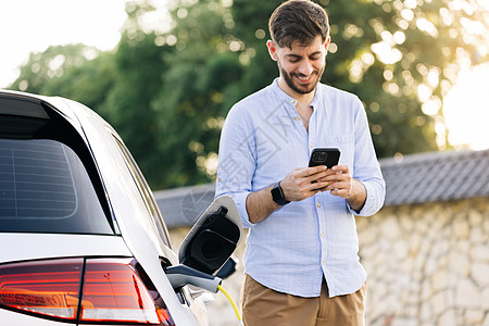 身穿蓝色衬衫 留着胡子的白人男子站在一辆正在充电的电动汽车旁边 并在智能手机上进行时间调整 从充电站为电动汽车充电图片