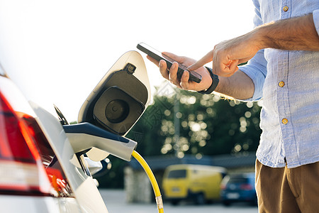 男性使用智能手机上的应用程序将电源线连接到电动汽车 从充电站插入电动汽车的人 商务人士在日落时在充电站给电动汽车充电图片