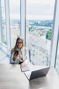 一位戴着眼镜 穿着时尚休闲服的金发开朗的欧洲女性正坐在一张桌子旁 拿着笔记本电脑 在做文书工作并打电话 办公室工作场所的商务女士图片
