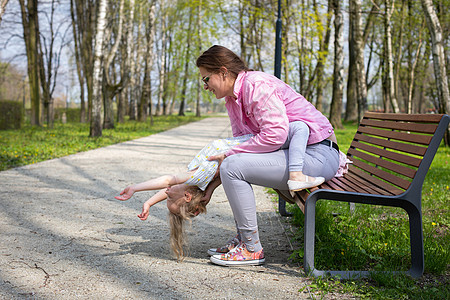 母亲和女儿坐在一个城市公园的长椅上玩体操拥抱儿童金发女郎时间喜悦眼睛低头黑发对方微笑图片