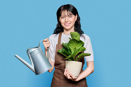 穿着围裙的微笑着年轻女子与饮用水浇灌植物的水罐 蓝底图片