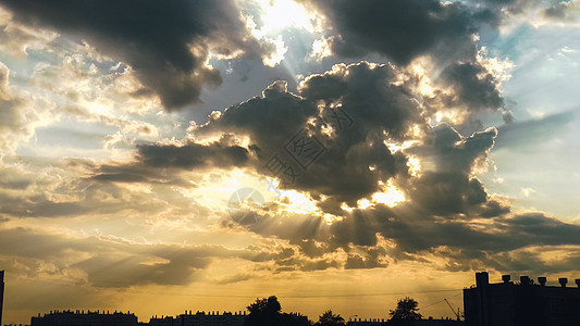 云层中的太阳将阳光投射在建筑物和树木轮廓上方的美丽天空上图片