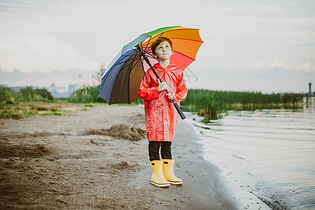 穿着红色雨衣和黄色橡胶靴的男孩站在河岸边 手里拿着彩虹伞 小学生站在秋天的湖边 在岸边穿着防水衣服的孩子公园寒意靴子气候海滩大衣图片