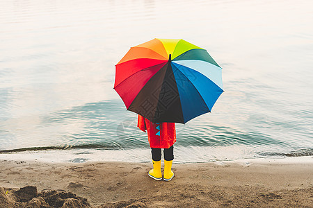 穿着红色雨衣和黄色橡胶靴的男孩拿着彩虹伞站在海滩上 带着五颜六色的雨伞看着大海的孩子 一件防水红色外套的学童拿着多色伞图片