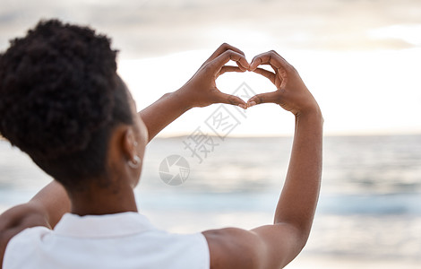 后视镜中一位非洲裔美国女性的手在户外的天空下做心形 日落时分 自信的黑人女性站在外面的海滩上 指着大海 健康与爱图片