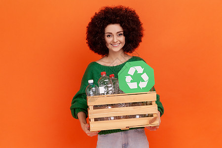 满意的非洲发型妇女穿着绿色随身毛衣 装满一箱空塑料瓶和绿色标志 回收利用 生态概念图片