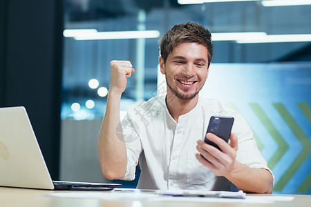 在办公室工作的快乐男人 看着电话屏幕看新闻 读喜讯报的好男人商业报酬拳头商务人士胜利企业家手机男性手势图片