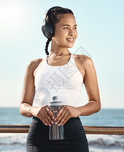 快乐的西班牙裔年轻女运动员一边戴着耳机听音乐 一边拿着水瓶微笑 一边在海滨长廊进行有氧运动 一边休息图片