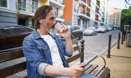 独自坐在长椅上的中年男子离开办公室拿着数字平板电脑 在老城区的街道上用无线耳机打电话喝外卖咖啡 在旅途中的业务概念图片