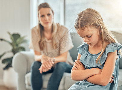 悲伤的白人小女孩坐在客厅里 双臂交叉 感到沮丧 而她的母亲则生气地看着 小可爱的女儿在家里让父母失望后感到不安图片