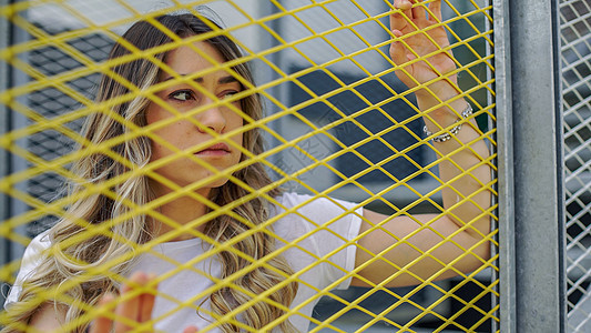 一名悲伤的年轻女性站在黄网围栏后面锁定监狱栅栏女士金属头发场地金发女孩迁移图片
