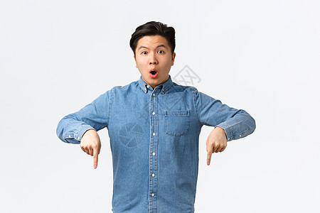 身穿蓝色衬衫的亚洲男人震惊而印象深刻 手指向下 无言地看着镜头 询问有关产品的问题 发现一些有趣的东西 站在白色背景图片