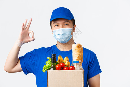 网上购物 食品配送和 covid-19 流行病概念 戴着医用面具 蓝色制服和手套的年轻帅气的亚洲男性快递员 保证 表现出好的姿态图片