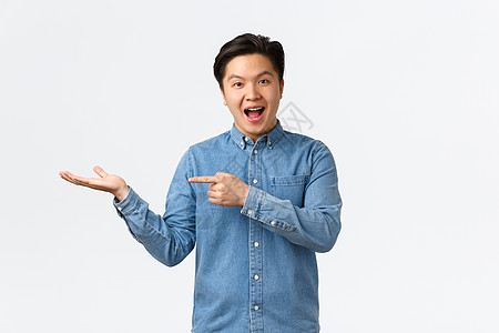 笑着开朗可爱的亚洲男人发布公告 展示手头的新产品 用手指指着广告的空白 站在白色背景上逗乐图片