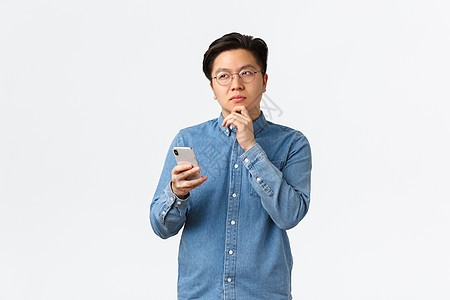 戴眼镜 有思想 有创造力的亚洲人在社交媒体上发帖时思考 目光移开 思考或做出决定 手持智能手机 在互联网上选择东西图片