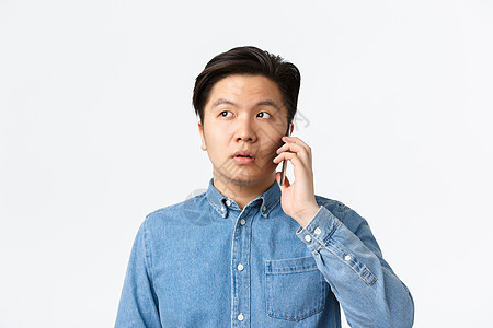 特写 优柔寡断的亚洲人在电话中发呆 交谈 困惑地看着别处 站在白色背景上 男人拿着智能手机靠近耳朵 点外卖图片