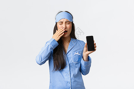 技术 人和家庭休闲理念 穿着睡面罩和睡衣的疲惫困倦的可爱亚洲女孩 打着哈欠疲惫地醒来 在智能手机或睡眠跟踪应用程序上显示时间图片