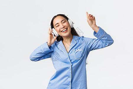 居家休闲 周末和生活方式的概念 穿着睡衣 无忧无虑 快乐的亚洲女孩享受着一天的假期 戴着耳机听音乐 脸上带着高兴的表情跳舞 站在图片