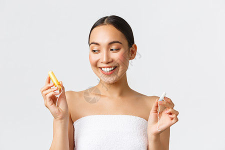 美容 个人和私密护理月经卫生概念 穿着浴巾 拿着卫生棉条的微笑可爱的亚洲女孩 看着一个带涂抹器的女孩 在白色背景下做出选择图片
