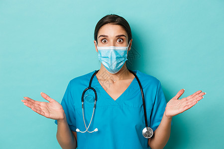 covid-19 的概念和检疫概念 惊讶的女医生特写 戴着医用口罩和磨砂膏 双手横开 耸耸肩 站在蓝色背景上图片