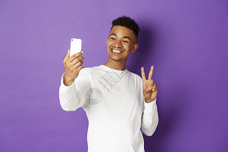 透过紫色背景站立 在手机摄影机上展示和平标志和微笑 男性博客情感技术手机快乐多样性发型运动衫成人互联网网络图片