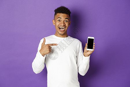 英俊而兴奋的非洲裔美国人 用手指指着智能手机屏幕 展示购物网站或应用程序 站在紫色背景上图片
