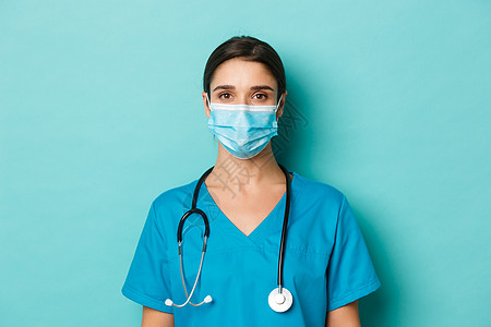 将年轻女医生用洗涤剂和医疗面具 看照相机 站在蓝底站立的结晶体和医学面罩特写起来背景图片