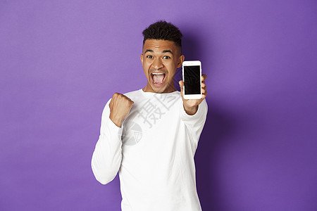 兴奋的非洲裔美国人展示手机屏幕 在网上赢得一些东西 为应用程序的成就而高兴 站在紫色背景上图片