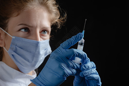 一位戴着防护面具和手套的女医生在黑色背景下拿着装有疫苗或注射剂的注射器 一名女护士准备在黑暗背景下进行注射 在黑色背景上独自手持图片