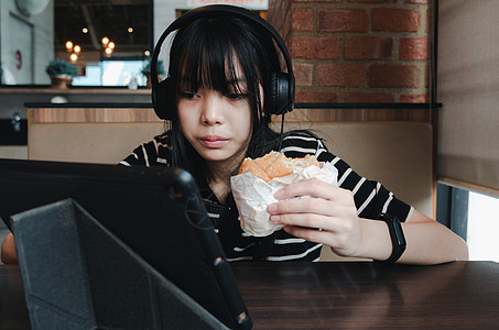 女孩在吃汉堡时要注意你的平板电脑 用耳机听音乐或使用社交媒体图片