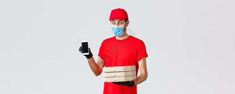 送餐 应用 网上杂货 非接触式购物和 covid19 概念 穿着红色制服 面罩和手套的搞笑快递员展示智能手机屏幕应用程序并拿着披图片