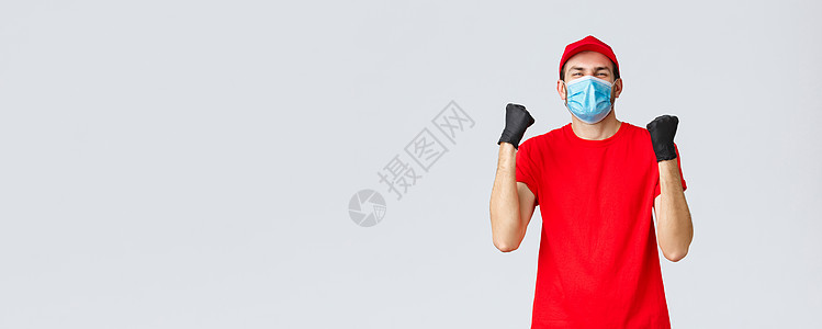 Covid19 自我隔离 网上购物和运输概念 身穿红色制服 戴着医用面罩 拳头抽动的兴奋 快乐的快递员欢欣鼓舞 送货员庆祝成功 图片
