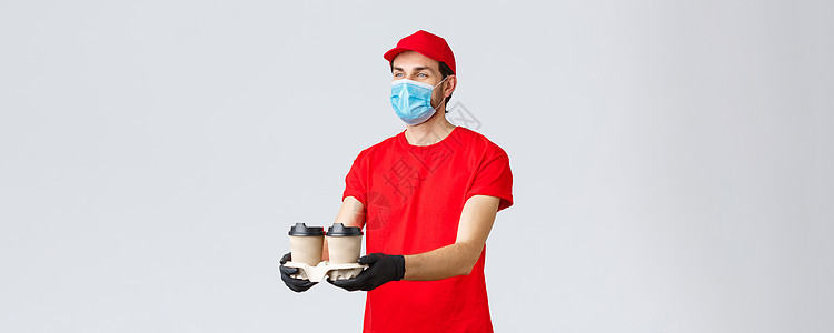 外卖 食品和杂货配送 covid19 非接触式订单概念 穿着红色制服 面罩和手套的愉快的快递员 给顾客发咖啡 站在灰色背景食物船图片