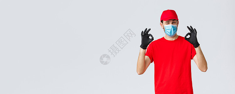 Covid19 自我隔离 在线购物和航运概念 穿红色帽子 T恤衫 戴防护面罩和冠状病毒手套的英俊送货员 表现好 同意和良好姿态邮图片