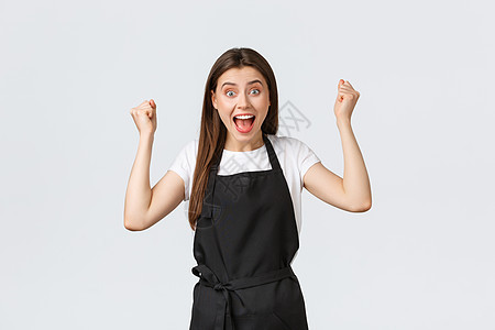 杂货店员工 小型企业和咖啡店的概念 身穿黑色围裙的快乐女咖啡师举起双手欢欣鼓舞 庆祝伟大的成就或成功图片