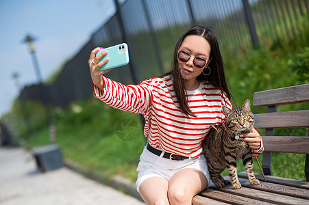 年轻女性坐在长椅上 跟一只小猫坐在一起 在户外的智能手机上拍自拍友谊猫咪宠物相机毛皮猫科动物后院胡须电话图片