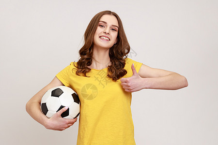 年轻漂亮的棕发少女拿着足球 带着露齿的微笑看着镜头 竖起大拇指 穿着休闲装图片