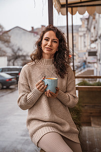 一个中年妇女穿着米色毛衣 手里拿着一个蓝杯子 在阳台的街头咖啡厅里草本植物女孩成人加热温度针织品茶碗餐厅女士羊毛图片